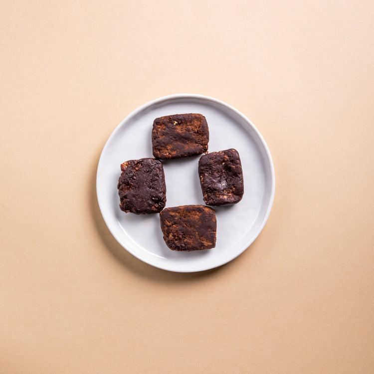 The image shows a simple white plate with four healthy cocoa fig bites. The brownies' fudgy texture and rich color stand out against a soft beige background, creating a minimalistic and appealing presentation.