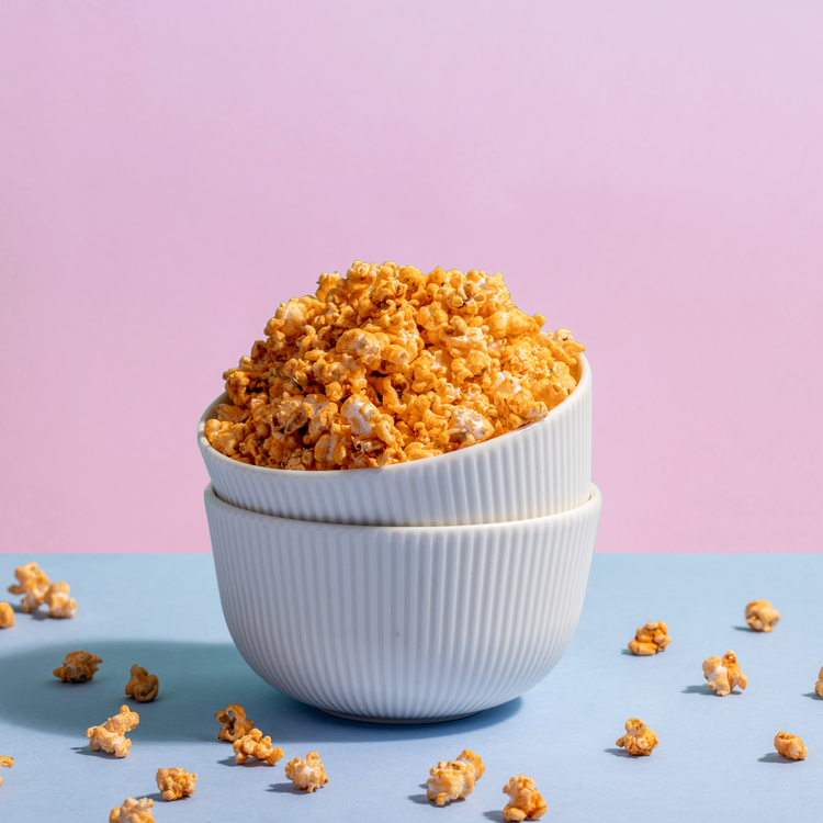 A stack of white ribbed bowls overflows with Barbeque Jowar Pops, set against a pink backdrop and scattered on a blue surface.