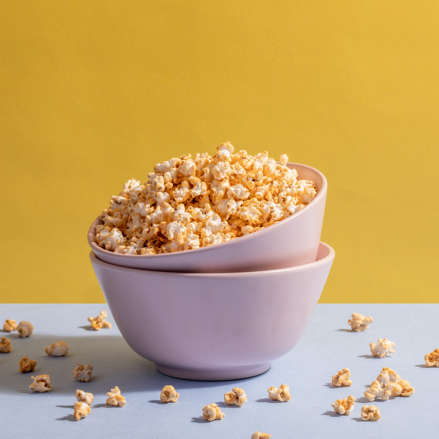 The image features two light pink bowls stacked together, filled with popped popcorn. The Chilli Cheese Jowar Pops is fluffy and golden, suggestive of fresh, buttery flavor. Surrounding the bowls are scattered pieces of popcorn on a soft blue surface. The background is a vibrant yellow, creating a cheerful and inviting atmosphere. The overall composition is bright and appetizing, perfect for a snack setting.