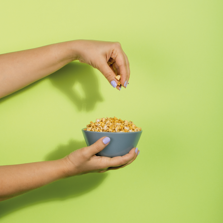 The image shows a person’s hands holding a light blue bowl filled with Chilli Cheese Jowar Pops against a vibrant green background. One hand is poised above the bowl, dropping a piece of popcorn into it. The composition evokes a playful and inviting atmosphere, highlighting the enjoyment of snack time. The purple nail polish on the fingers adds a touch of color to the scene.