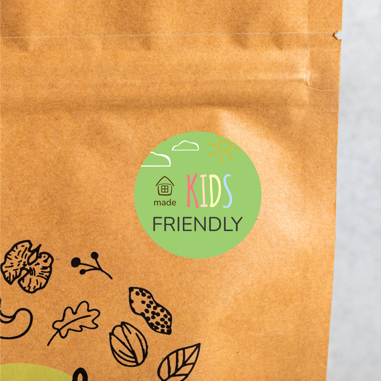 The image features a brown kraft Four Thirty packaging bag with a prominent circular sticker that reads "KIDS FRIENDLY." The sticker has a light green background, playful lettering in various colors, and small illustrations of a house, clouds, and the sun. Surrounding the sticker are simple black line drawings of leaves, nuts, and seeds, indicating the natural ingredients within. The overall design conveys a wholesome, inviting, and child-friendly appeal.