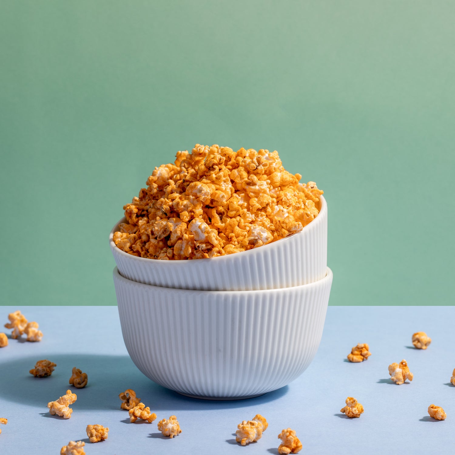 A stack of white ribbed bowls overflows with Peri Jowar Pops against a light green backdrop, scattered on a soft blue surface.