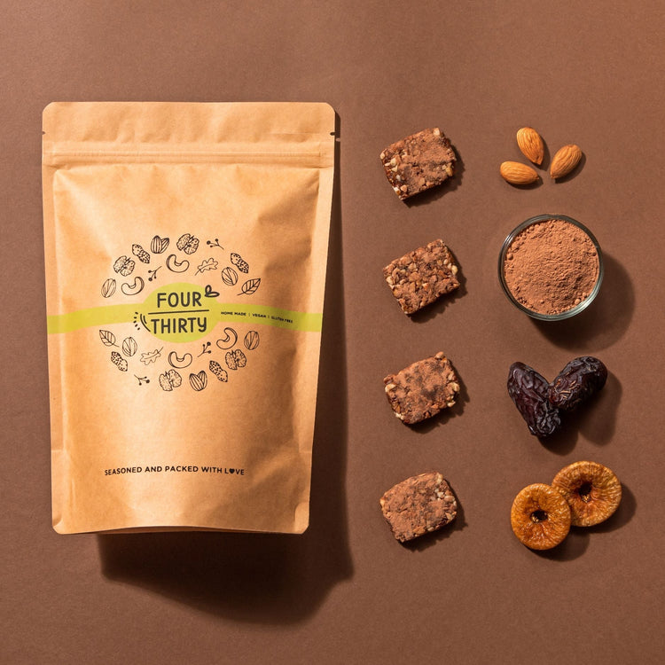 The image showcases a brown kraft package labeled "FOUR THIRTY," featuring illustrations of nuts and seeds. It highlights being homemade, vegan, and gluten-free. Next to the package are three textured energy bars, a small glass bowl of brown powder, whole almonds, dried figs, and a heart-shaped date, emphasizing a wholesome and natural approach to nutrition.